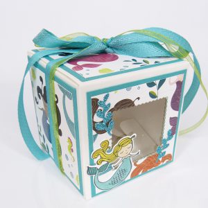 handdekorierte Schachtel mit Meerjungfrau-Motiv, Geschenkbox mit Fenster und Schleife, maritime Geschenkverpackung mit Glitzerdetails, Schachtel mit türkisfarbenem Band, Nahaufnahme Meerjungfrau auf Geschenkbox, Unterwasser-Deko auf Schachtel, Geschenkbox für Kindergeburtstag mit Meerjungfrau