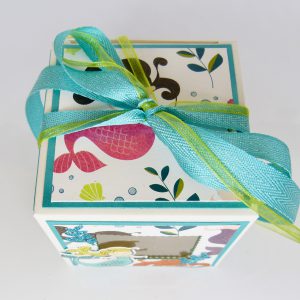 handdekorierte Schachtel mit Meerjungfrau-Motiv, Geschenkbox mit Fenster und Schleife, maritime Geschenkverpackung mit Glitzerdetails, Schachtel mit türkisfarbenem Band, Nahaufnahme Meerjungfrau auf Geschenkbox, Unterwasser-Deko auf Schachtel, Geschenkbox für Kindergeburtstag mit Meerjungfrau