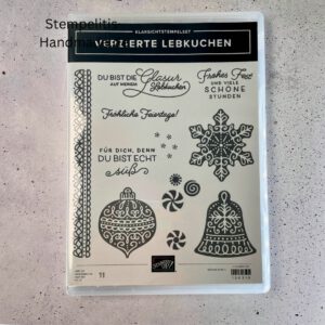 Bastelmaterialien für weihnachtliche Karten oder Projekte. Zu sehen sind grünes und rotes Tonpapier, gemustertes Designerpapier, ein Stempelset namens „Verzierte Lebkuchen“ mit passenden deutschen Sprüchen, metallische Stanzformen für Weihnachtsmotive (Stern, Glocke, Schneeflocke, Kugel), rote und weiße Kordel, goldene Zierdeckchen sowie Briefumschläge