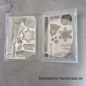 Bastelmaterialien für weihnachtliche Karten oder Projekte. Zu sehen sind grünes und rotes Tonpapier, gemustertes Designerpapier, ein Stempelset namens „Verzierte Lebkuchen“ mit passenden deutschen Sprüchen, metallische Stanzformen für Weihnachtsmotive (Stern, Glocke, Schneeflocke, Kugel), rote und weiße Kordel, goldene Zierdeckchen sowie Briefumschläge