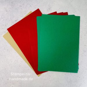 Bastelmaterialien für weihnachtliche Karten oder Projekte. Zu sehen sind grünes und rotes Tonpapier, gemustertes Designerpapier, ein Stempelset namens „Verzierte Lebkuchen“ mit passenden deutschen Sprüchen, metallische Stanzformen für Weihnachtsmotive (Stern, Glocke, Schneeflocke, Kugel), rote und weiße Kordel, goldene Zierdeckchen sowie Briefumschläge