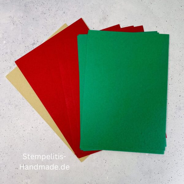 Bastelmaterialien für weihnachtliche Karten oder Projekte. Zu sehen sind grünes und rotes Tonpapier, gemustertes Designerpapier, ein Stempelset namens „Verzierte Lebkuchen“ mit passenden deutschen Sprüchen, metallische Stanzformen für Weihnachtsmotive (Stern, Glocke, Schneeflocke, Kugel), rote und weiße Kordel, goldene Zierdeckchen sowie Briefumschläge