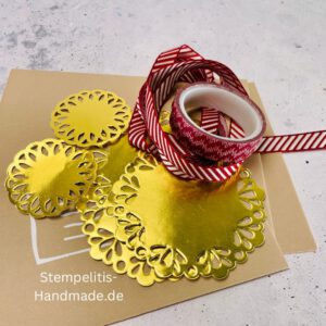 Bastelmaterialien für weihnachtliche Karten oder Projekte. Zu sehen sind grünes und rotes Tonpapier, gemustertes Designerpapier, ein Stempelset namens „Verzierte Lebkuchen“ mit passenden deutschen Sprüchen, metallische Stanzformen für Weihnachtsmotive (Stern, Glocke, Schneeflocke, Kugel), rote und weiße Kordel, goldene Zierdeckchen sowie Briefumschläge