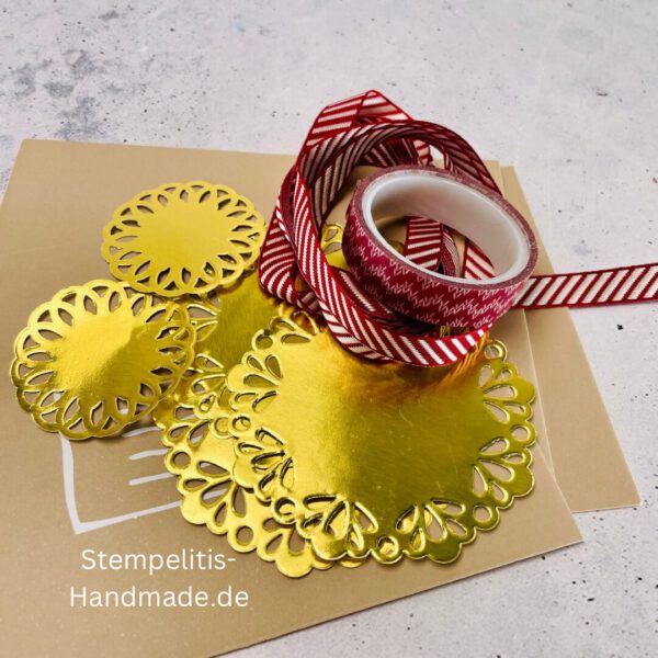 Bastelmaterialien für weihnachtliche Karten oder Projekte. Zu sehen sind grünes und rotes Tonpapier, gemustertes Designerpapier, ein Stempelset namens „Verzierte Lebkuchen“ mit passenden deutschen Sprüchen, metallische Stanzformen für Weihnachtsmotive (Stern, Glocke, Schneeflocke, Kugel), rote und weiße Kordel, goldene Zierdeckchen sowie Briefumschläge